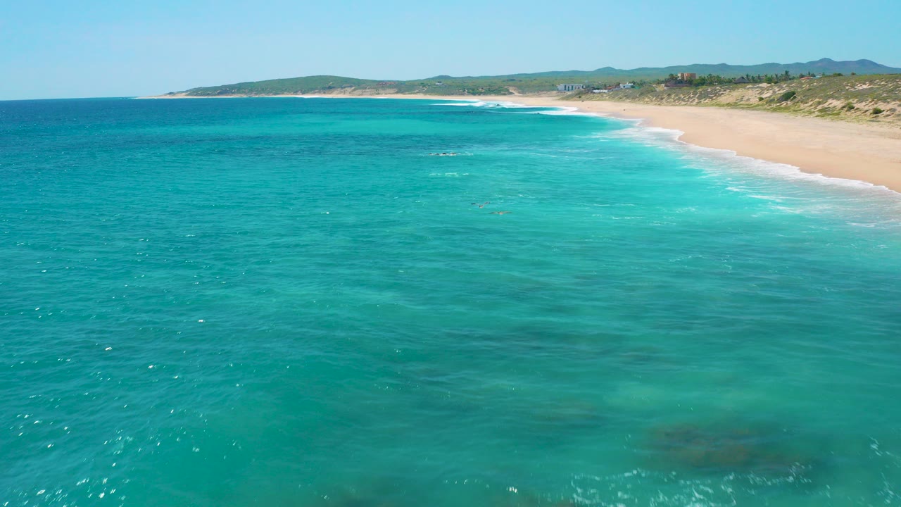 Aerial frame over clear turquoise water at El Cardoncito, Baja California Sur, with a sandy shoreline to the right, low dunes beyond, distant hills on the horizon, and no visible people.