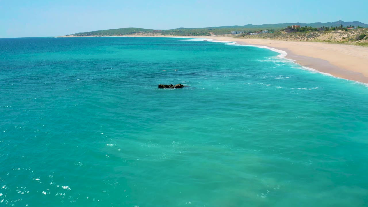 Aerial frame over clear blue water at El Cardoncito, with the rock outcrop slightly below center, pale sand sweeping along the upper right, low vegetation behind the beach, and no visible people.