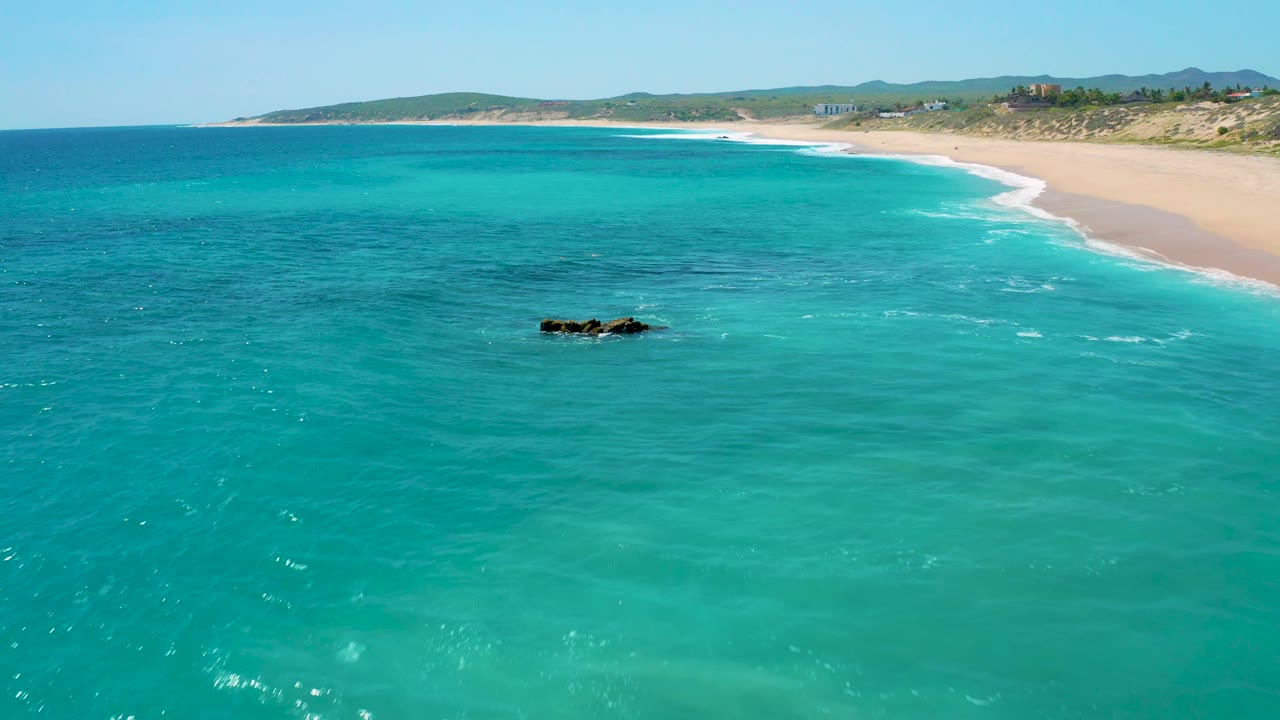 El Cardoncito shoreline seen from above, with shallow turquoise water across the frame, dark rocks nearer the right side, a pale beach at the top, distant low hills, and no visible people.