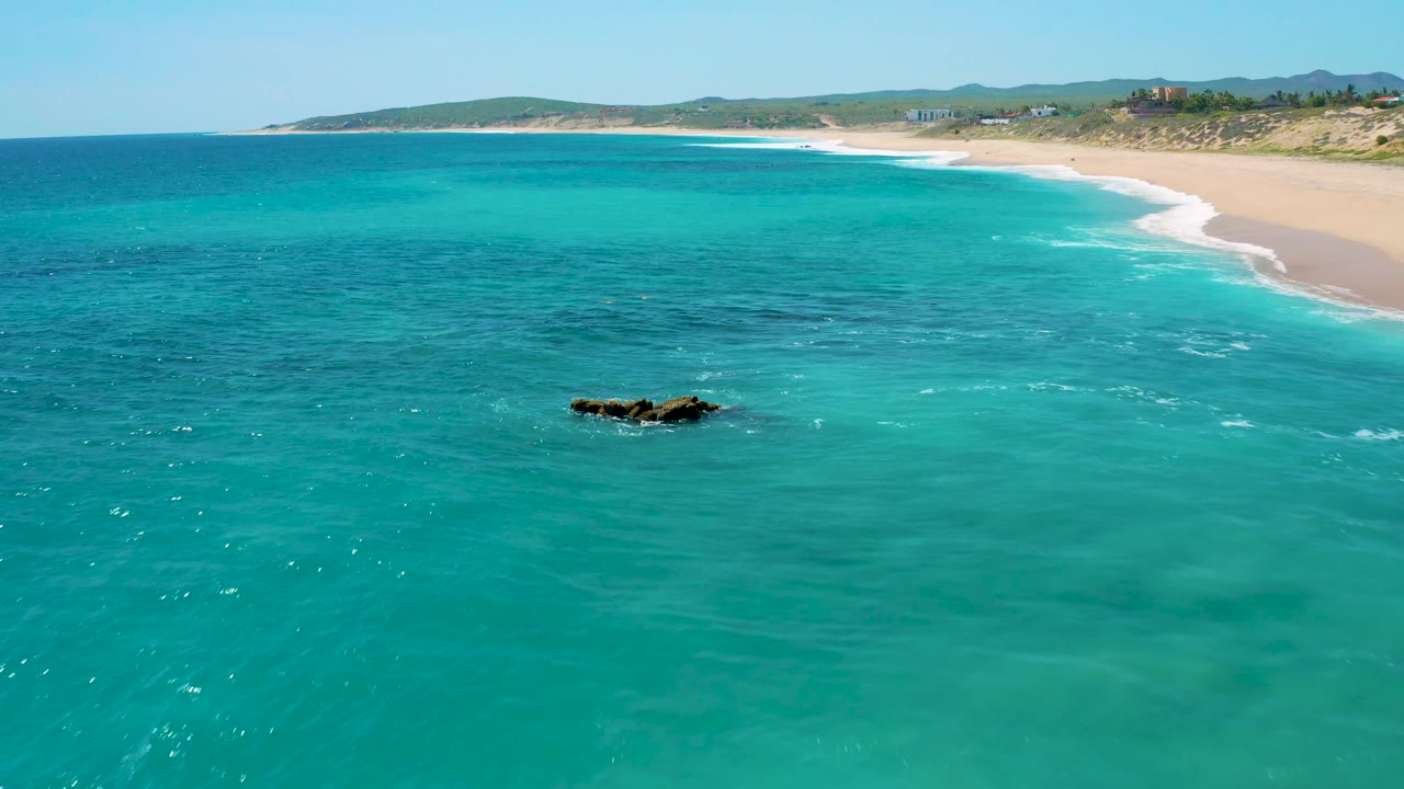 Clear water fills the lower frame at El Cardoncito, with a dark rock outcrop left of center, a curving sandy shoreline across the top, low scrub beyond the beach, and no visible people.