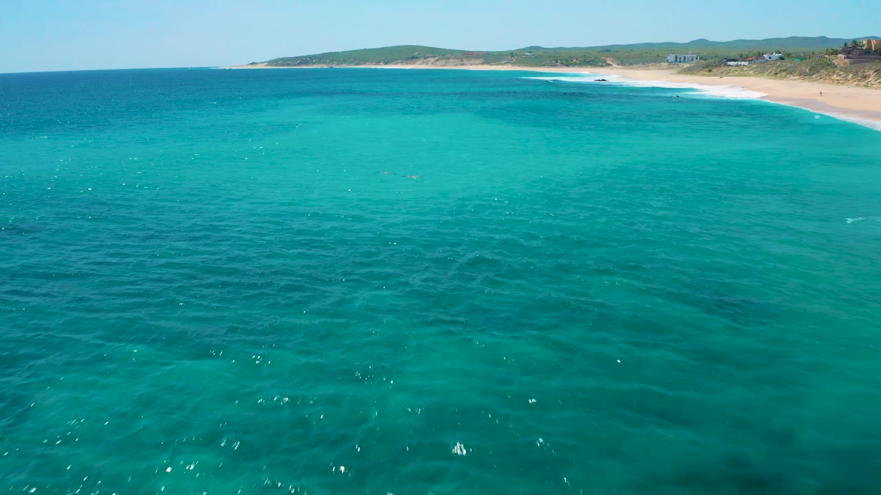 The drone view looks across clear blue-green water toward the El Cardoncito beach, with subtle wave marks, coastal hills, and no visible people.