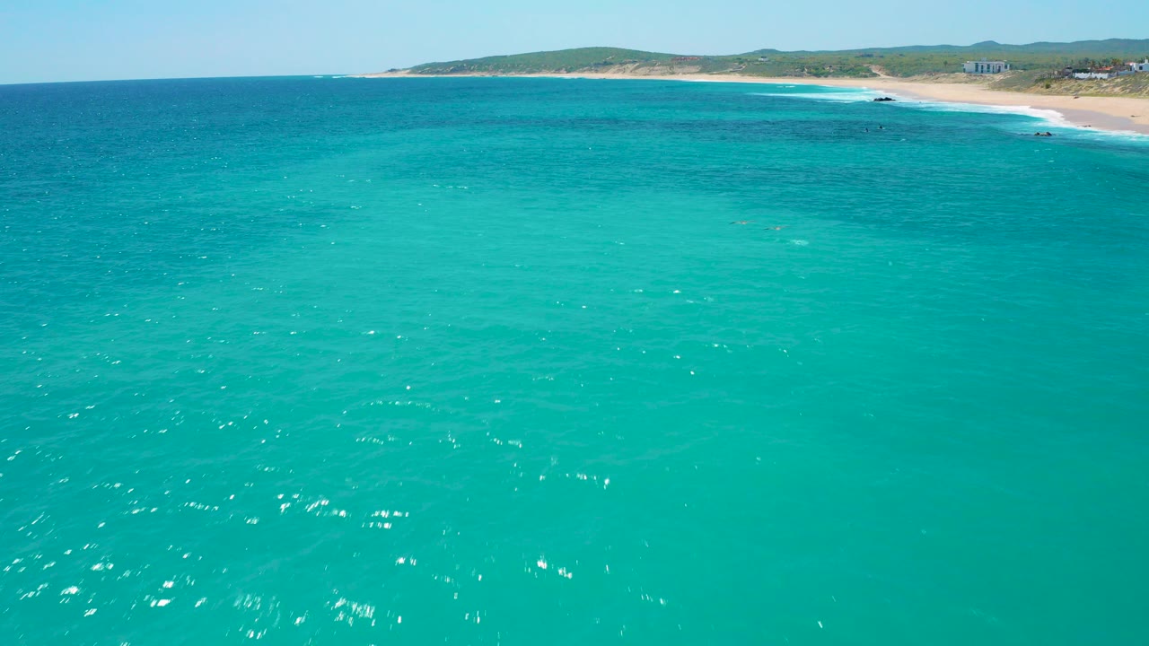Wide aerial view over turquoise water toward the El Cardoncito shoreline in Baja California Sur, with low hills beyond the sand and no visible people.