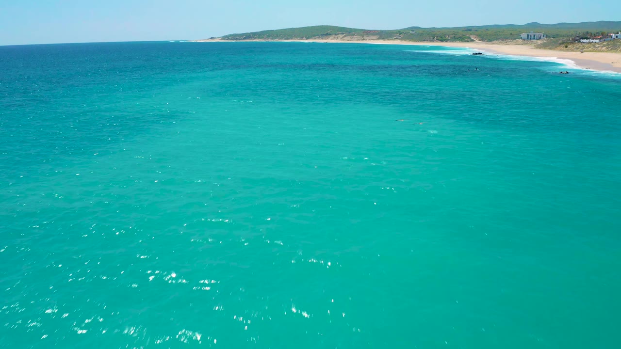 Aerial frame over rippled blue-green ocean at El Cardoncito, with darker patches under the water and a thin surf line on the distant sandy beach, no visible people.
