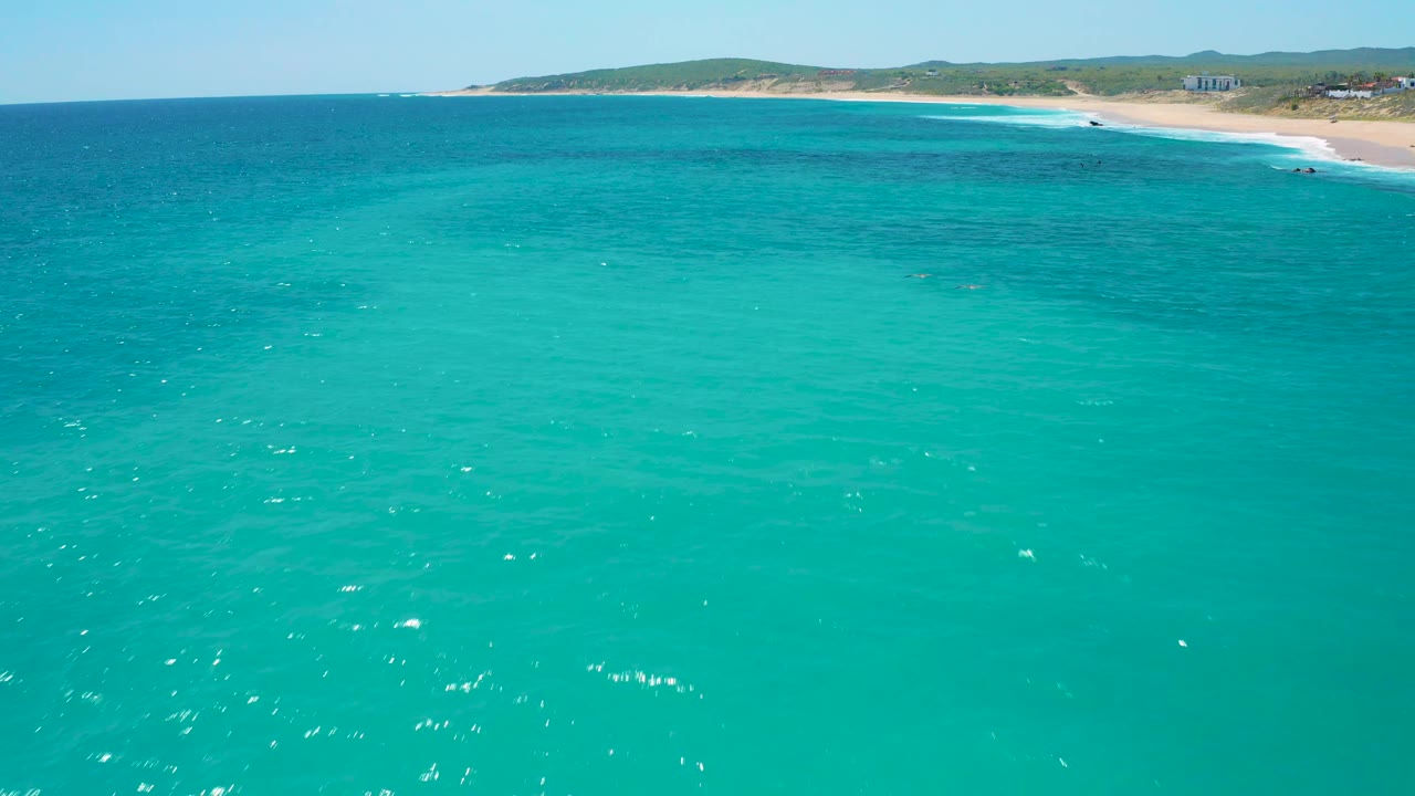 High aerial view of turquoise water leading toward the El Cardoncito beach, with small white wave breaks and low green hills behind the shore, no visible people.