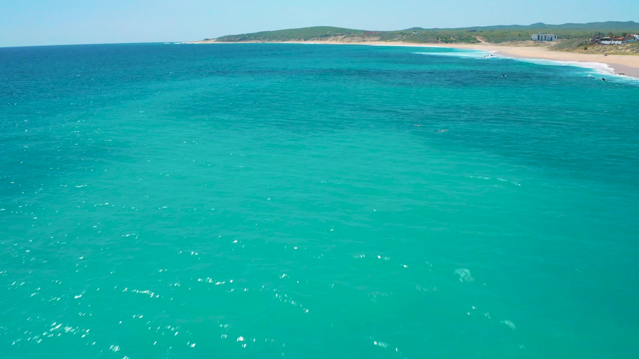 Offshore aerial view at El Cardoncito showing clear turquoise water in the foreground, pale surf curling onto sand, and a low developed coastline in the distance, no visible people.