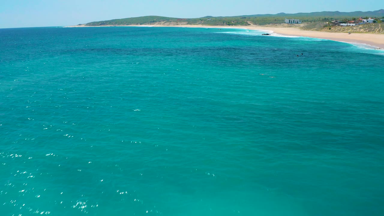 Aerial frame over broad teal water with the sandy beach compressed near the top and low Baja California Sur hills beyond, with no visible people.