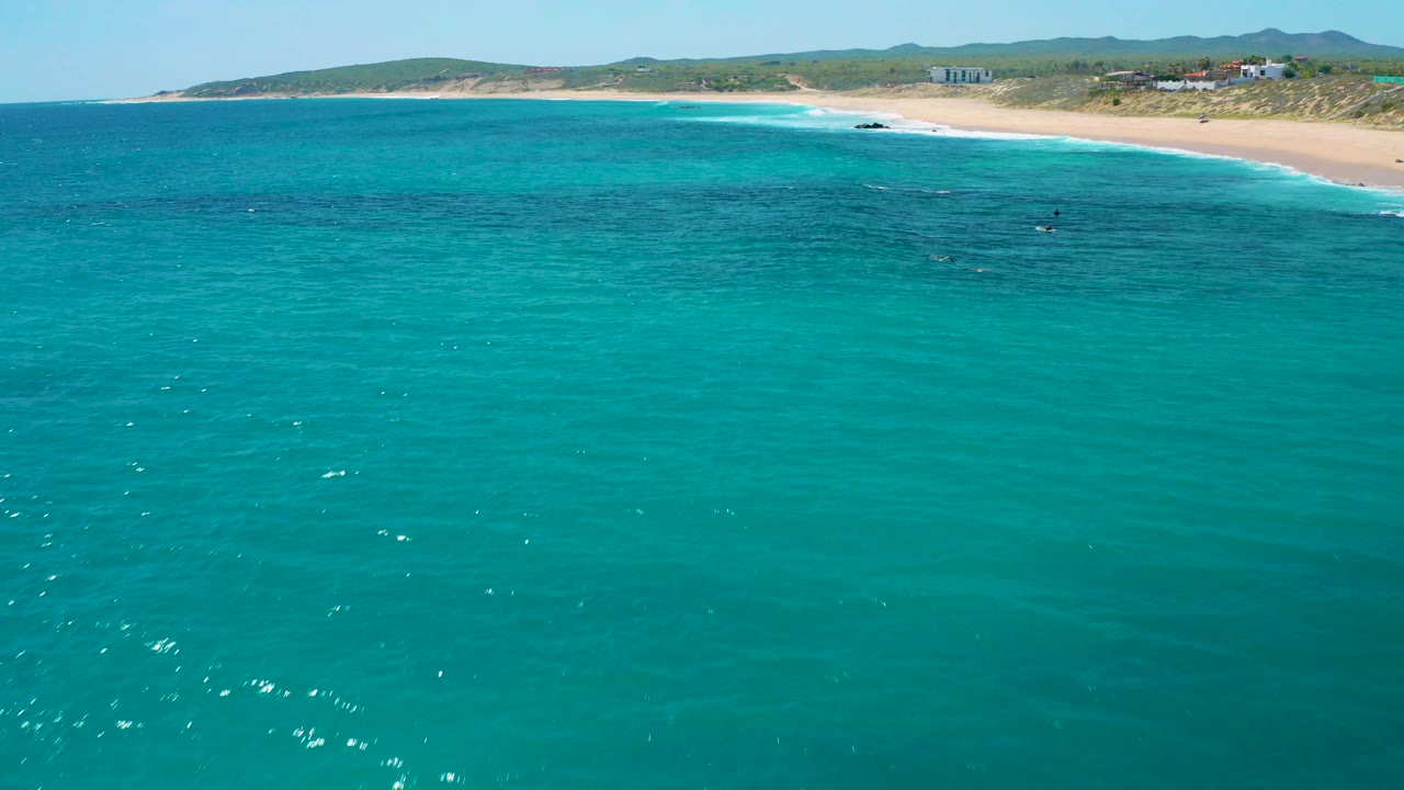 The coastline sits higher in frame with turquoise shallows, a white surf band, sandy dunes, and low hills behind, with no visible people.