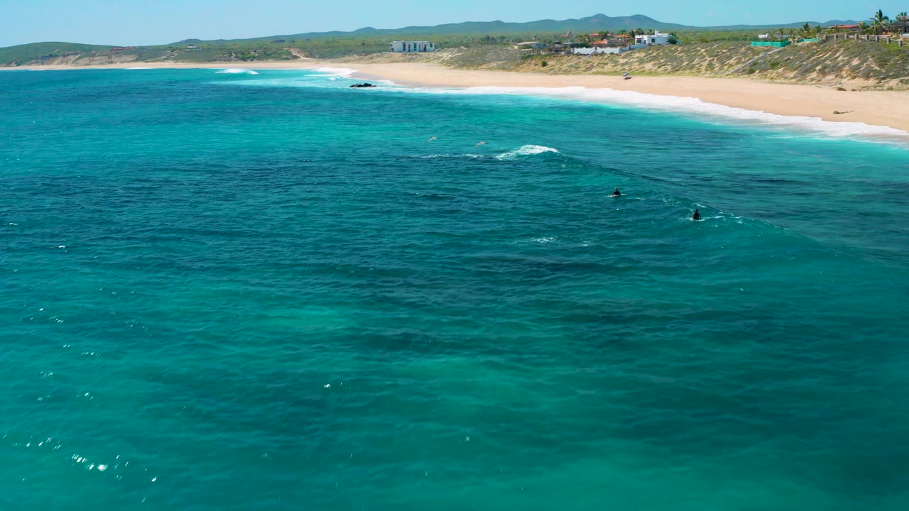 Green and turquoise water fills the foreground at El Cardoncito, with small white wave breaks, pale sand, low dunes, distant buildings, and no visible people.
