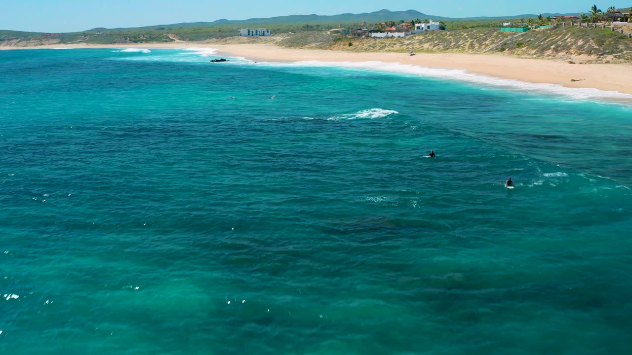 Wide aerial view at El Cardoncito showing dark underwater patches in green water, a narrow surf line, sandy beach, low dunes, and no visible people.