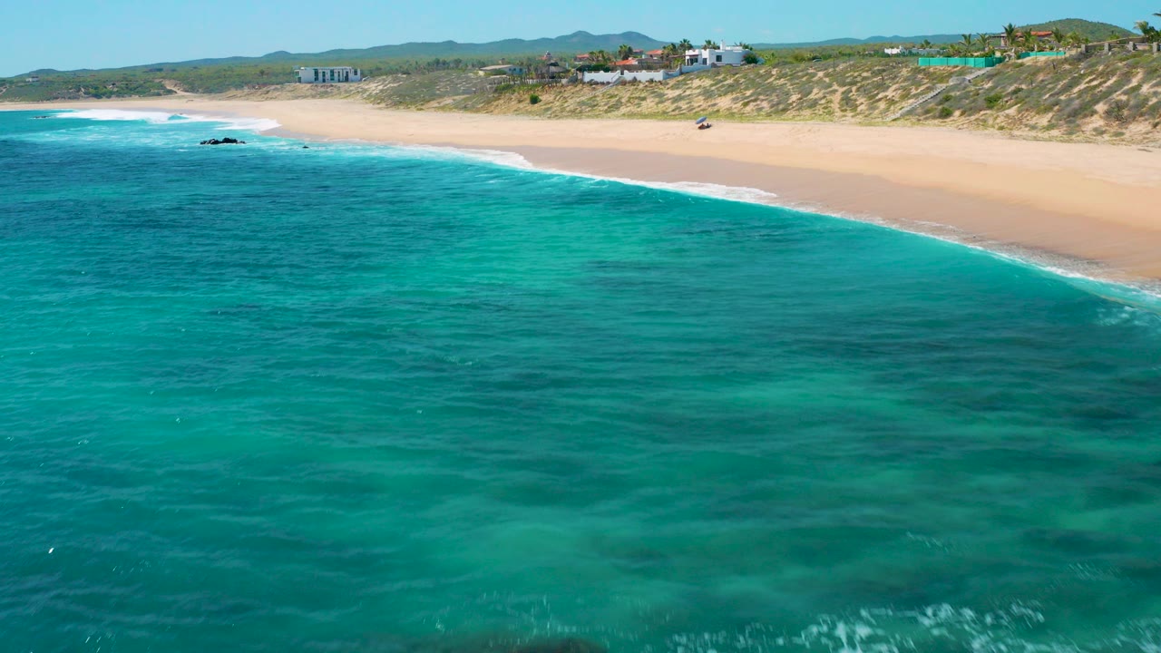 Aerial view over turquoise water toward the sandy beach at El Cardoncito, Baja California Sur, with low dunes, distant structures, and no visible people.