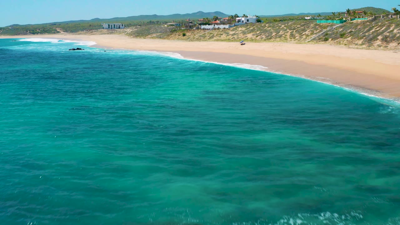 Wide beach aerial with aqua ocean, pale sand, dunes, and distant buildings.