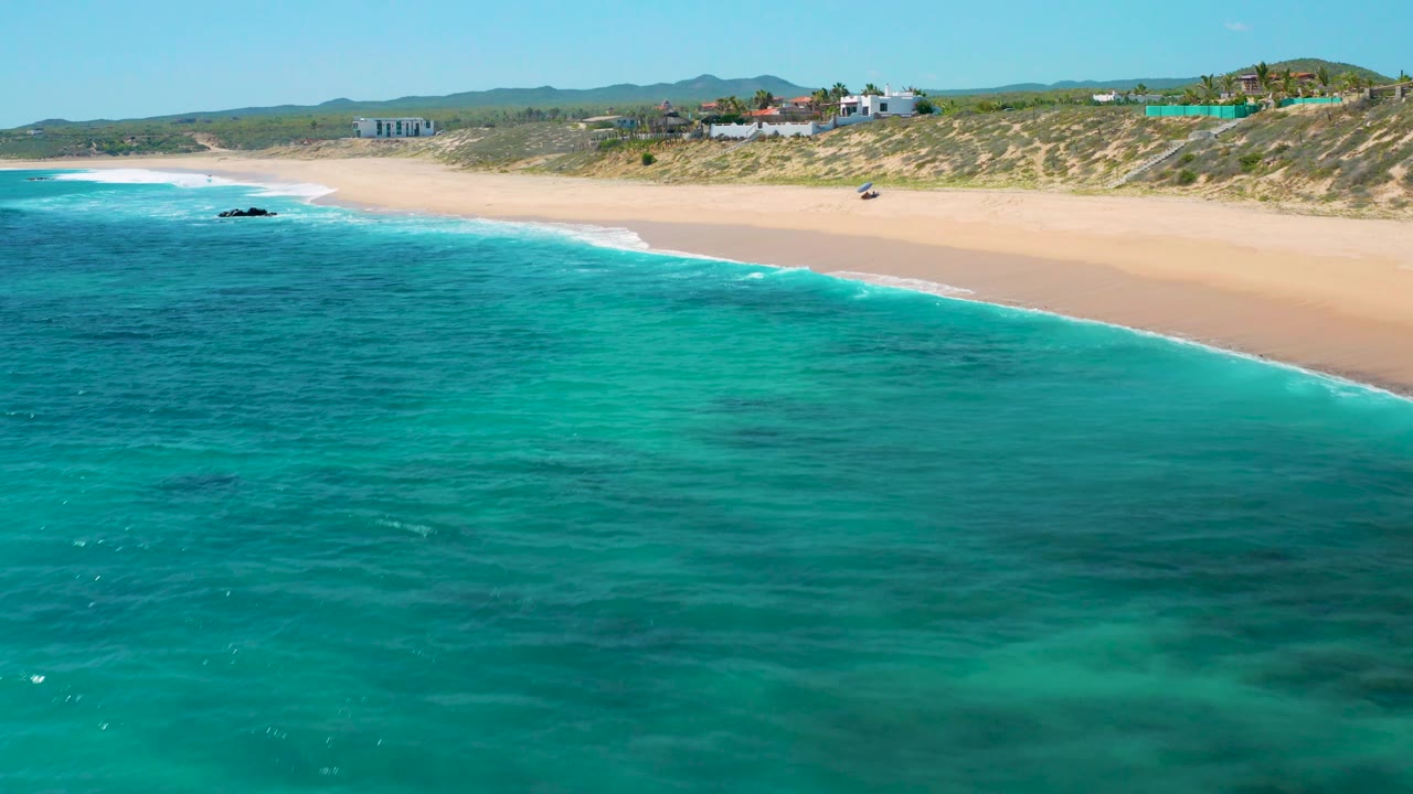 Aerial coastline view in El Cardoncito with turquoise water across the foreground, a curving sand beach, dunes, beachfront structures, and no visible people.