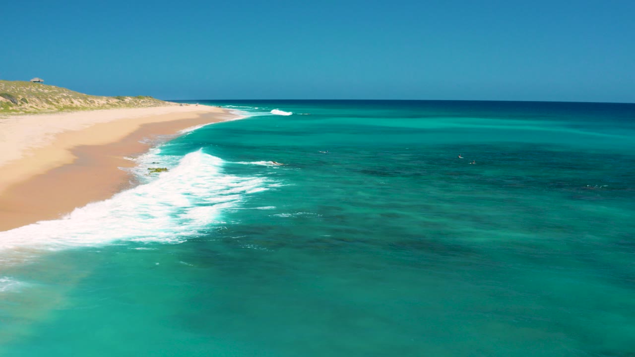 Aerial view at El Cardoncito, Baja California Sur, with pale sand on the left, white surf entering clear teal water, low dunes beyond the beach, and no visible people.