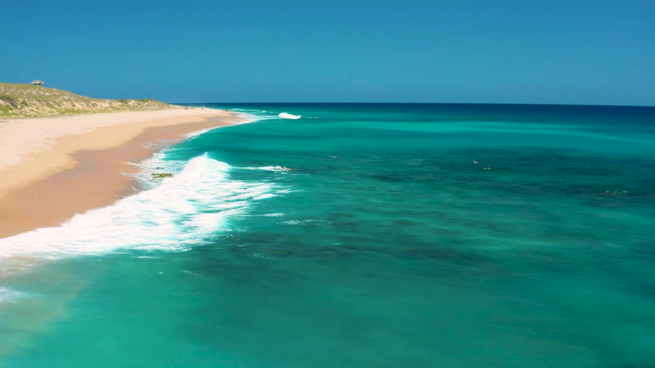 Teal water and white waves curve along an empty sandy shoreline.