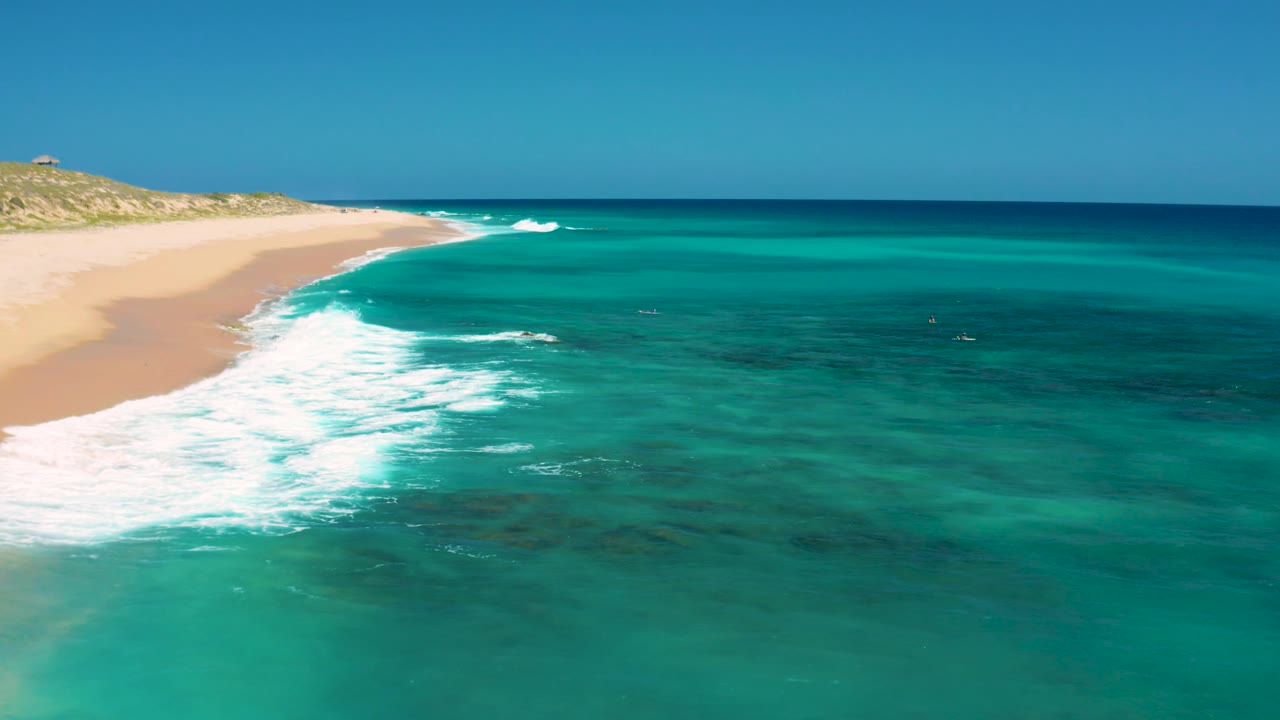 At 0:02, the aerial view shows more open water across the frame, with the sandy El Cardoncito beach receding at left, a small breaker near shore, and no visible people.