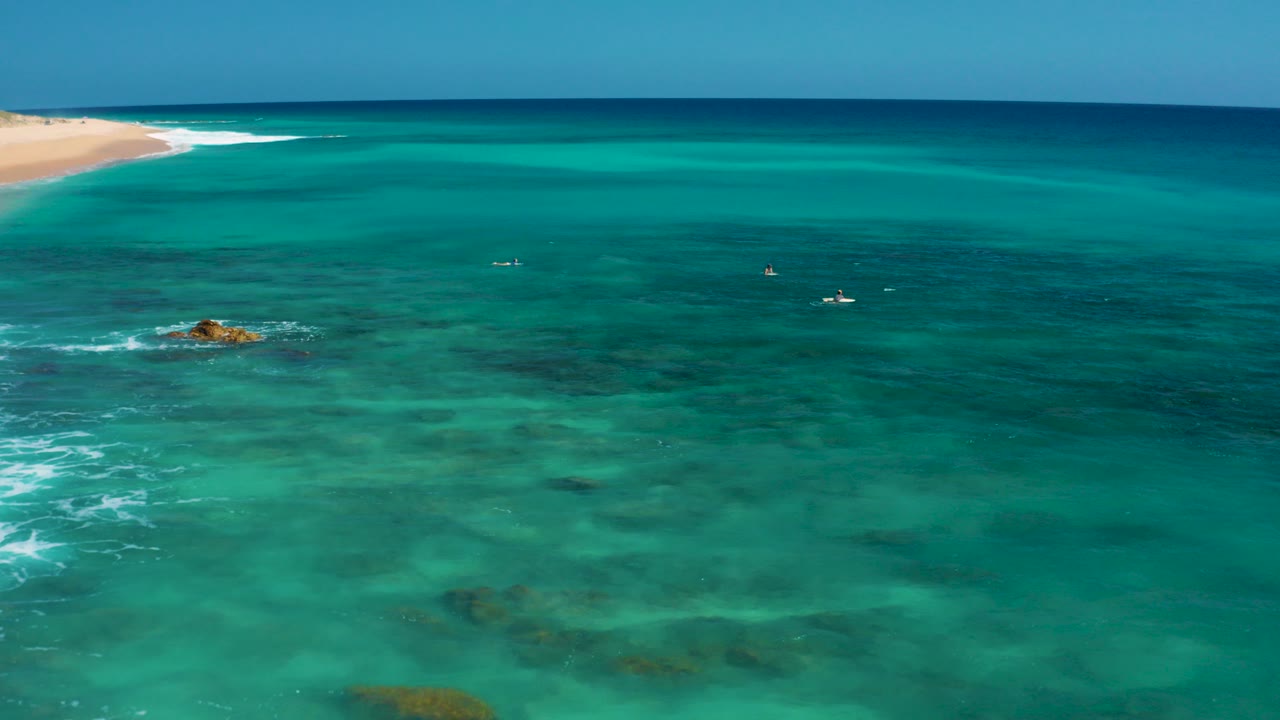 Aerial view over clear turquoise water at El Cardoncito, Baja California Sur, with reef patches below, a small rock near the left side, distant sand at the upper left, and no visible people.