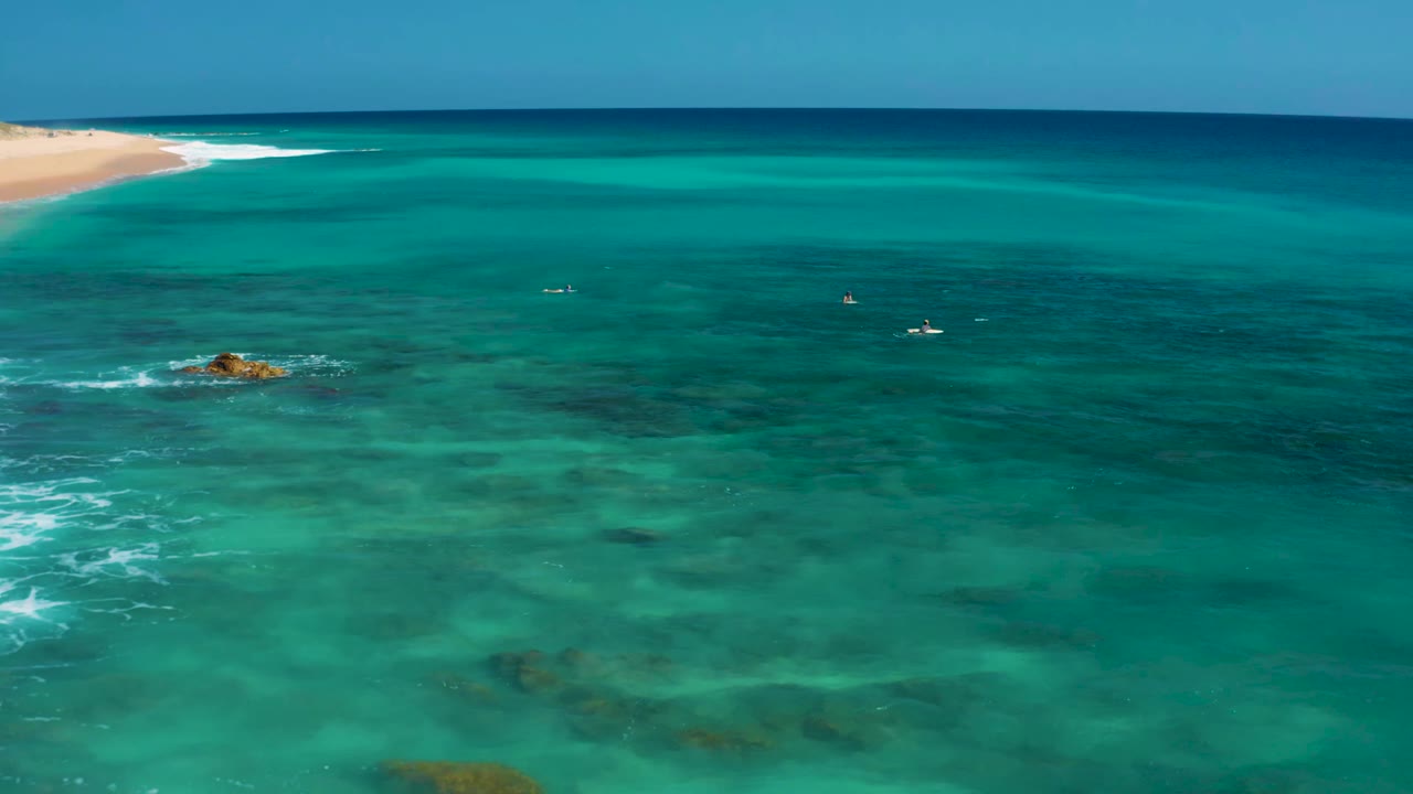 Aerial frame at El Cardoncito showing clear blue-green water, dark reef shapes beneath the surface, a small rock left of center, distant sandy shore, and no visible people.
