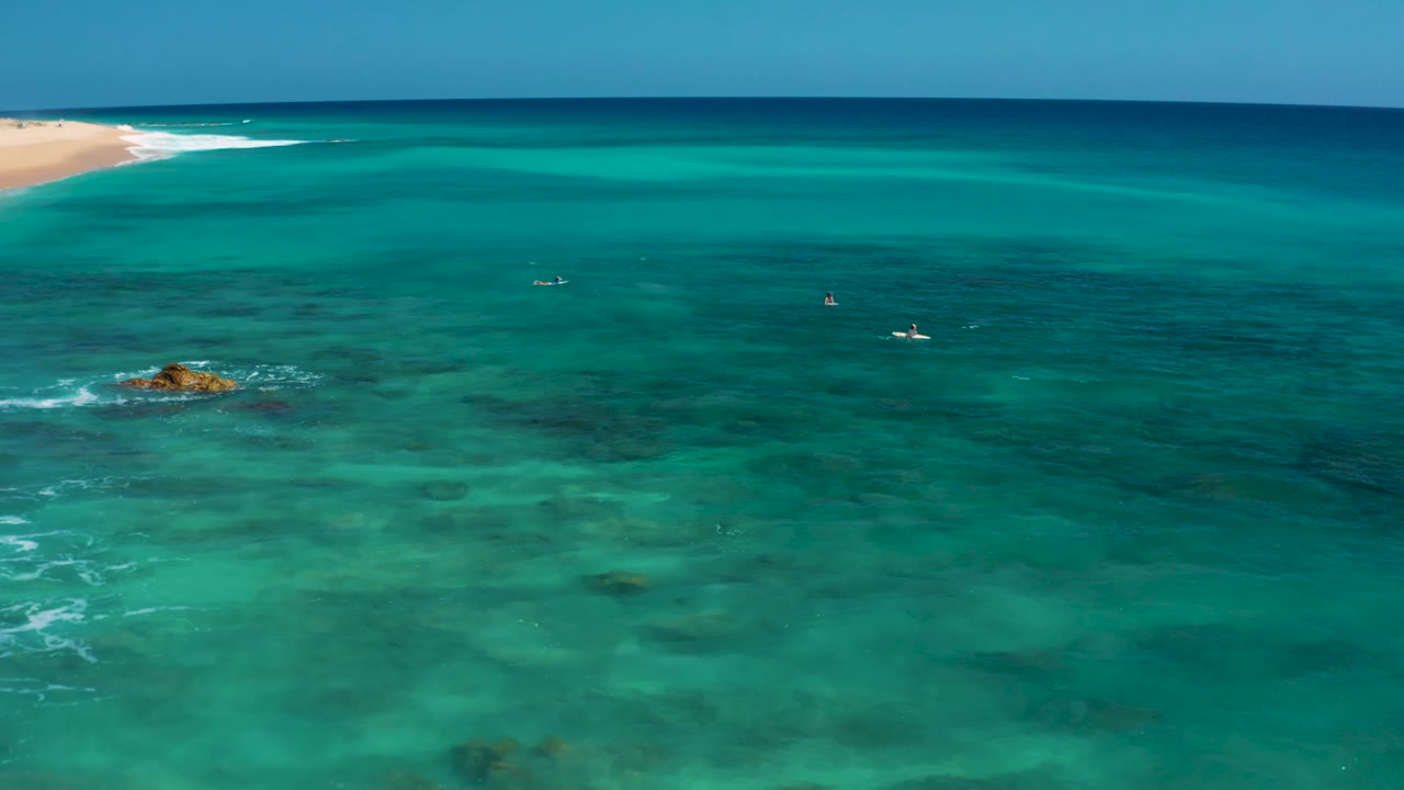 Wide aerial view over El Cardoncito water with the sandy shoreline clipped at the upper left, a small rock in the shallows, scattered reef patches, and no visible people.