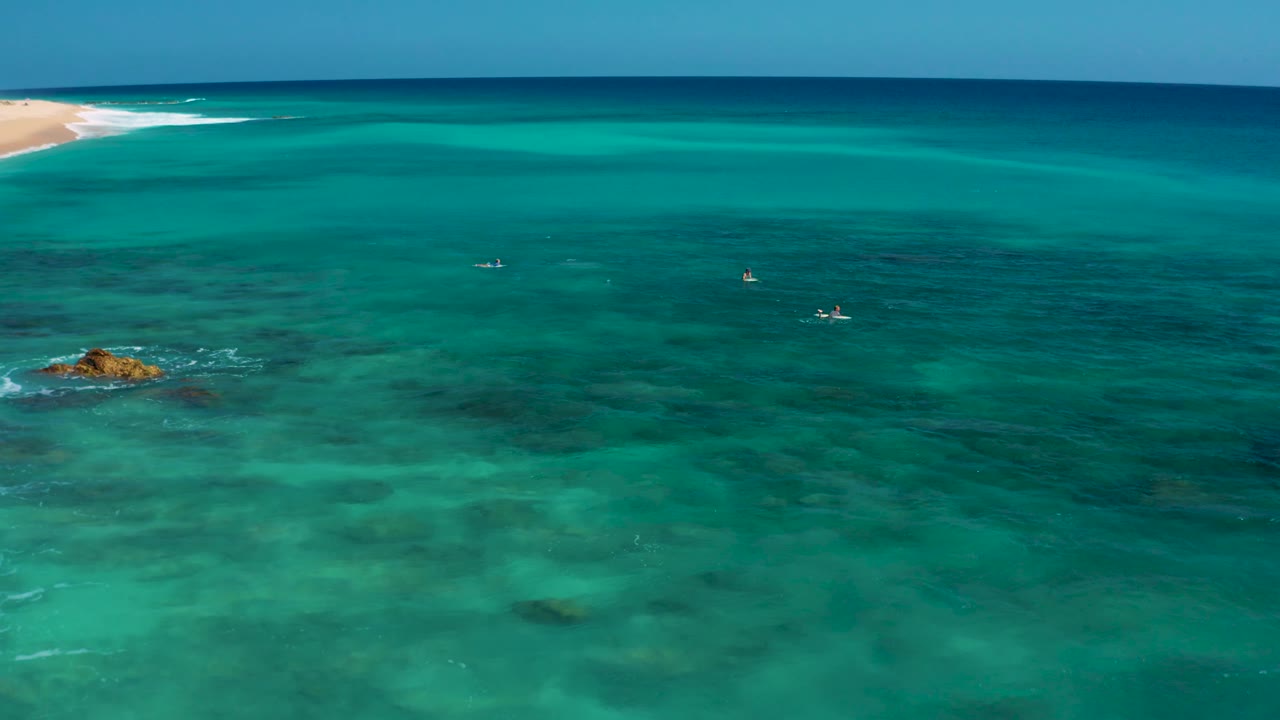 Aerial frame over El Cardoncito clear water, with the small exposed rock lower left, reef texture spreading through the foreground, distant shoreline at upper left, and no visible people.