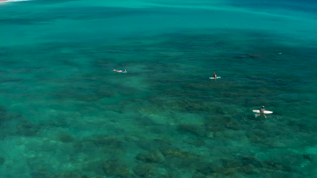 At 0:02, three surfers on boards are visible from above in El Cardoncito, surrounded by blue-green water and irregular dark reef patches.