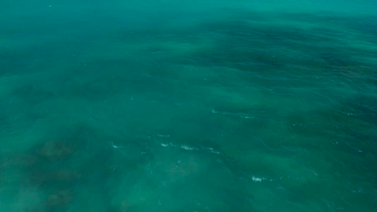 Overhead ocean frame from El Cardoncito showing teal water, soft ripple lines, and broad darker areas beneath the surface with no visible people.