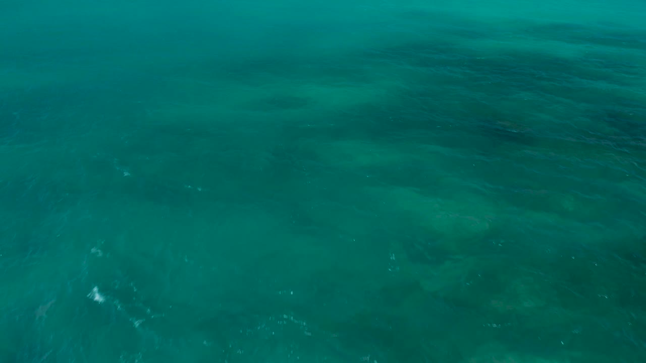 Turquoise ocean seen from above in El Cardoncito, with mottled darker seabed forms across the center and no visible people.