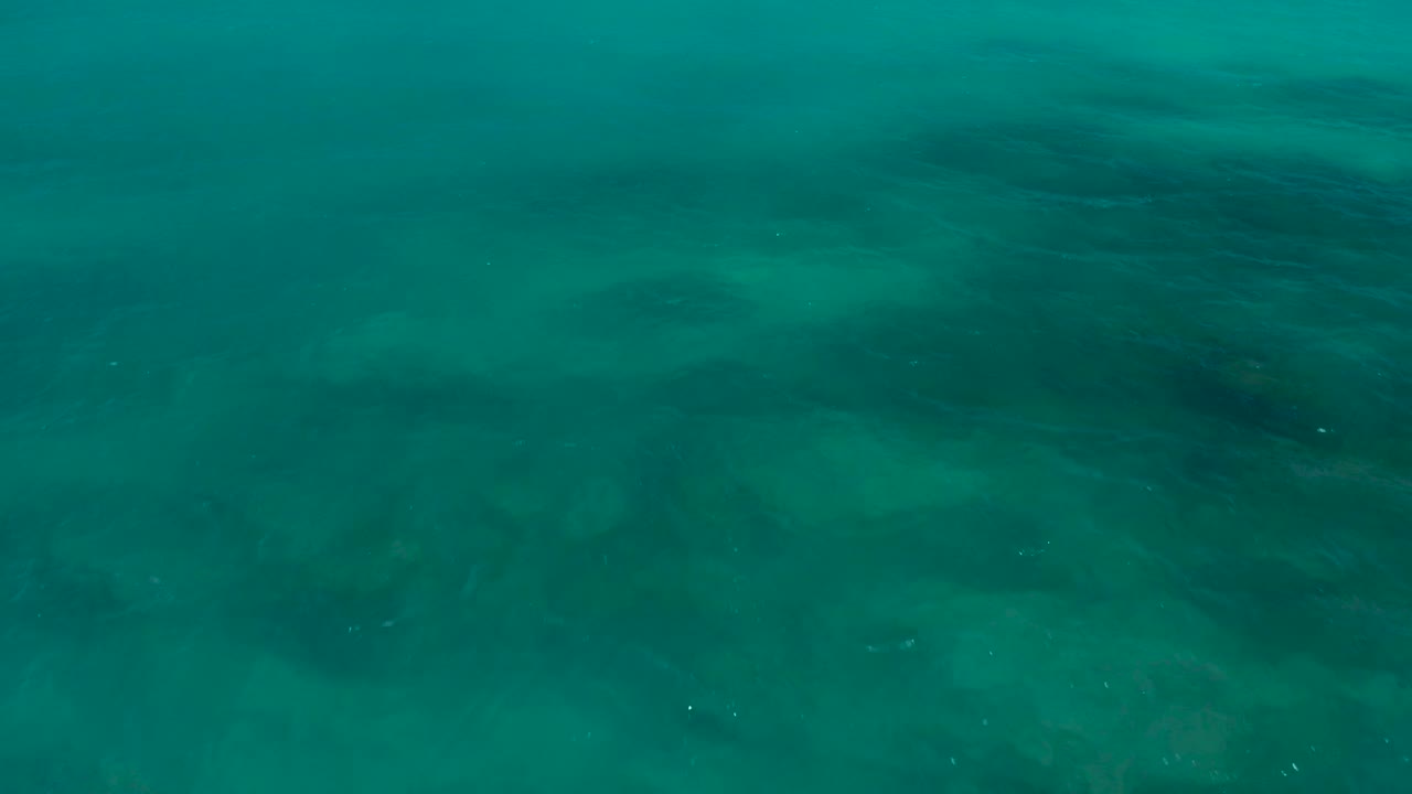 Aerial view of clear turquoise water near El Cardoncito, with a tiny bright speck near the lower left edge and no visible people.