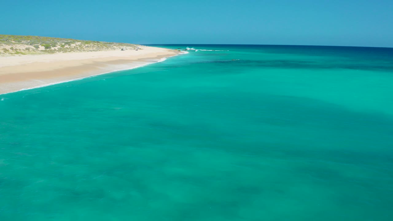 Aerial view over El Cardoncito, Baja California Sur, with turquoise water filling the frame, pale sand at upper left, low dune vegetation beyond it, and no visible people.