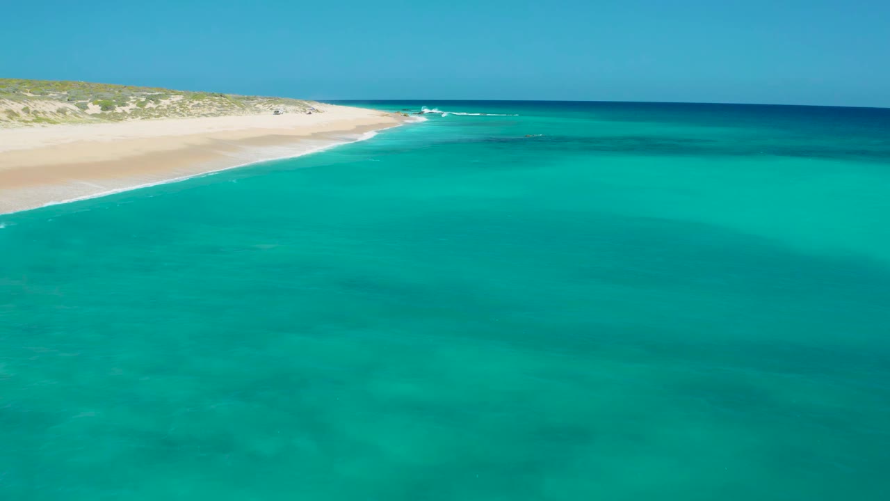 Aerial frame at El Cardoncito showing a wider spread of blue-green water, the sandy shoreline curving from the upper left, low coastal vegetation, and no visible people.