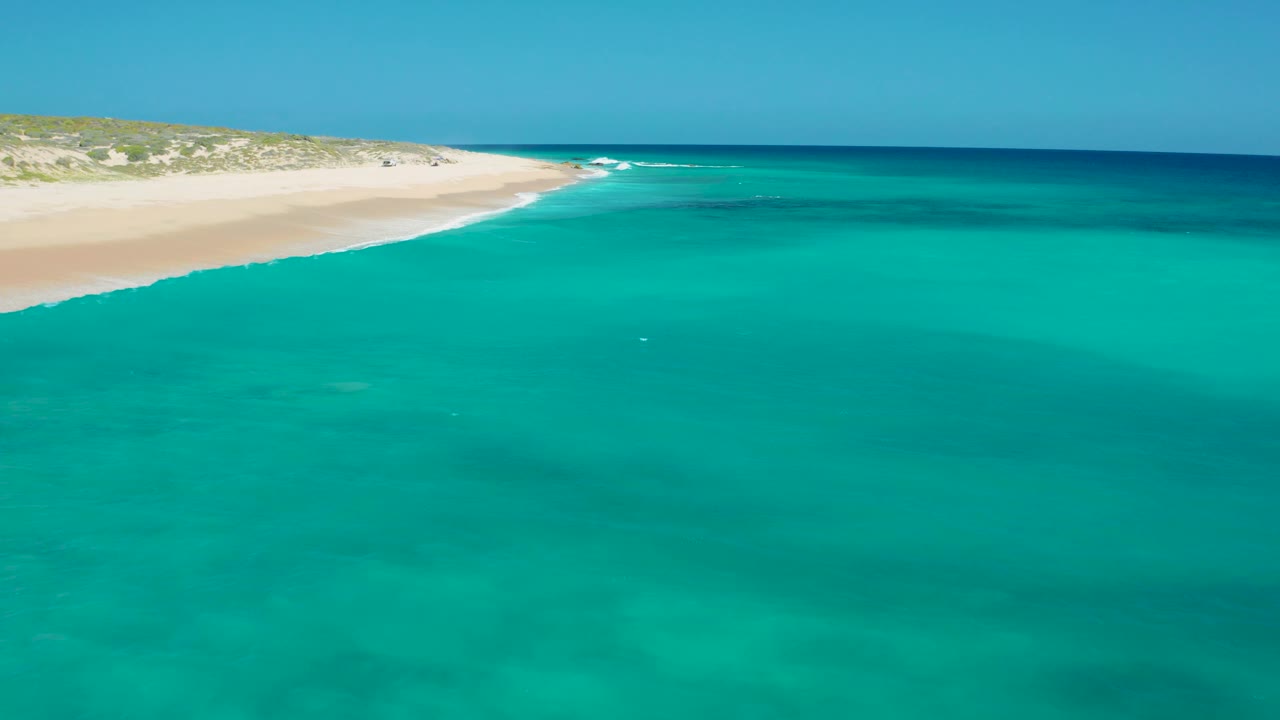Aerial view of El Cardoncito with broad turquoise water in front, a pale beach strip at upper left, low vegetation behind the sand, a dark offshore band, and no visible people.