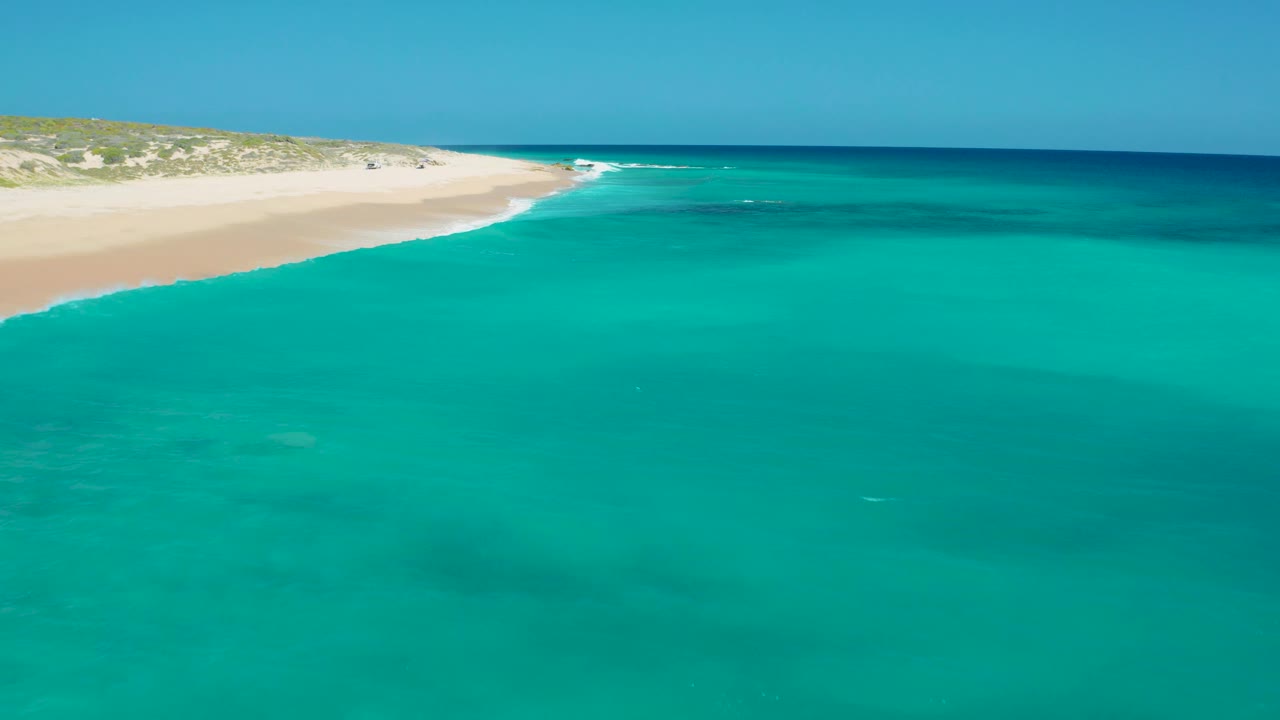 El Cardoncito shoreline seen from above, with small white surf touching the pale sand, clear teal water across the foreground, low dunes in back, and no visible people.