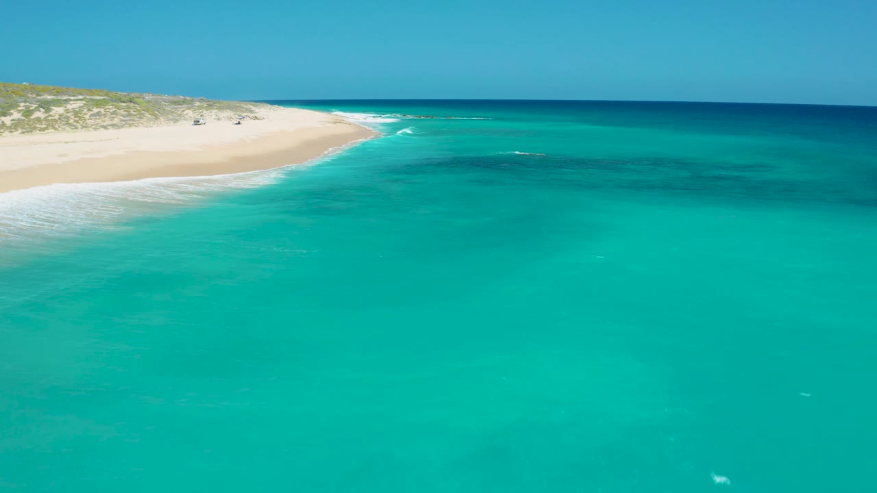 A wide aerial frame shows shallow turquoise water leading to a white sandy El Cardoncito shoreline, low scrub behind it, and no visible people.