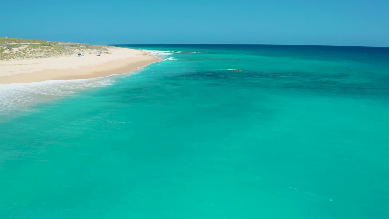 Foam from gentle waves runs along the left shoreline at El Cardoncito, with bright turquoise water across the frame and no visible people.
