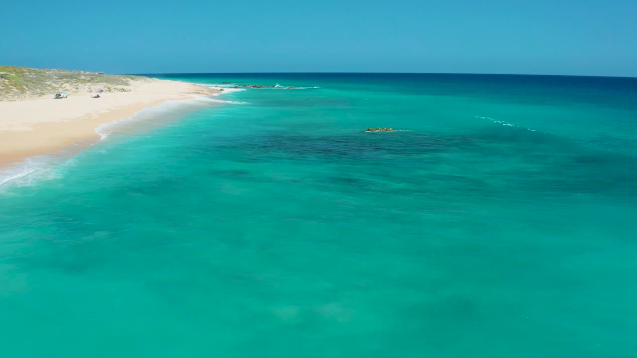 Aerial coastal frame from El Cardoncito showing clear turquoise water across the foreground, sandy shoreline at left, submerged dark reef areas, small rock outcrops, and no visible people.