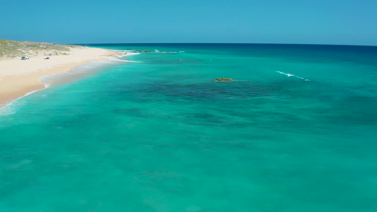 High aerial view at El Cardoncito with the pale sand beach receding along the left side, open turquoise sea to the right, dark underwater patches near center, and no visible people.