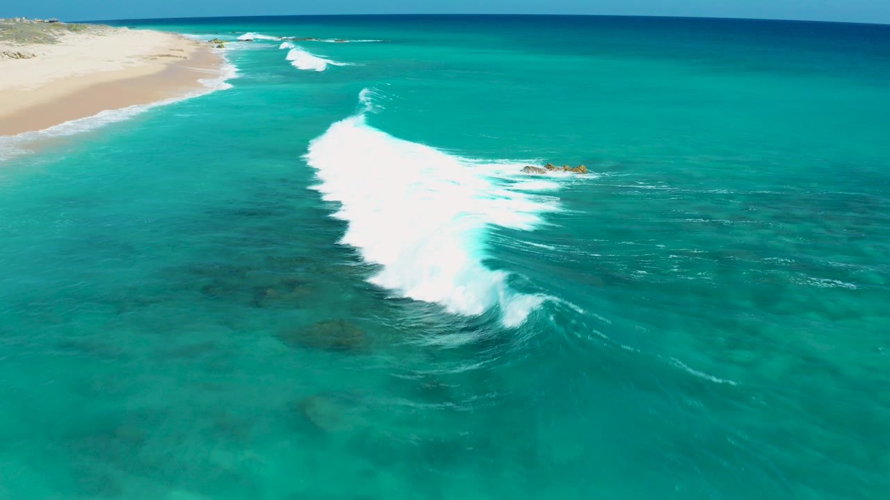 Aerial frame over El Cardoncito, Baja California Sur showing turquoise water, a pale sandy shoreline, white surf breaking diagonally, small rocks offshore, and no visible people.