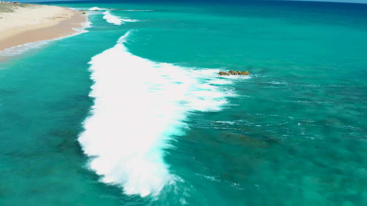 Aerial frame over El Cardoncito, Baja California Sur showing a larger white breaker spreading near small exposed rocks, with bright turquoise water and no visible people.