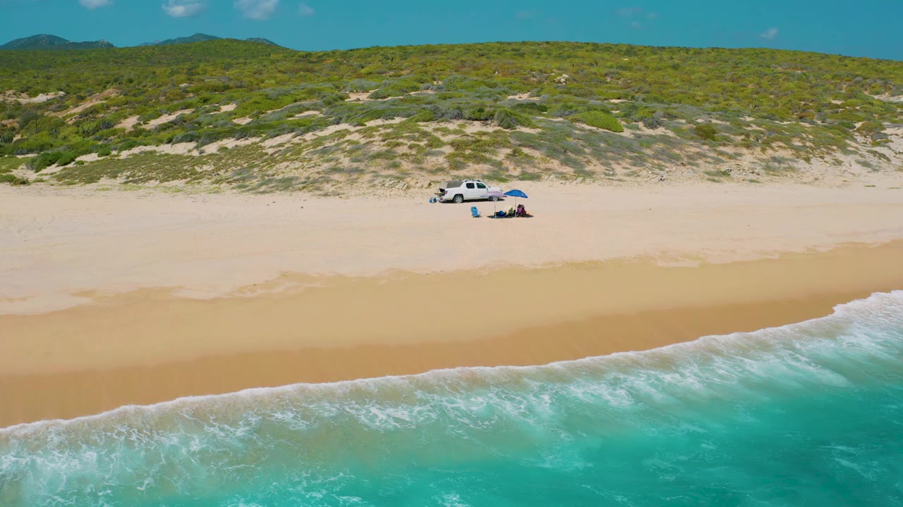 Wide aerial frame at El Cardoncito showing the white truck and blue umbrella set back from the turquoise shoreline, with low dune vegetation and no visible people.