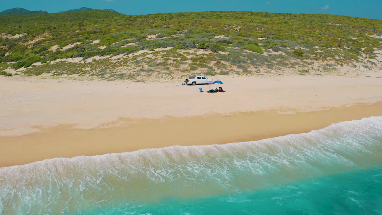 Aerial view of El Cardoncito with a foamy wave spreading along the lower edge, a broad empty sand band, dune scrub behind it, and the white truck setup small in the upper middle, with no visible people.