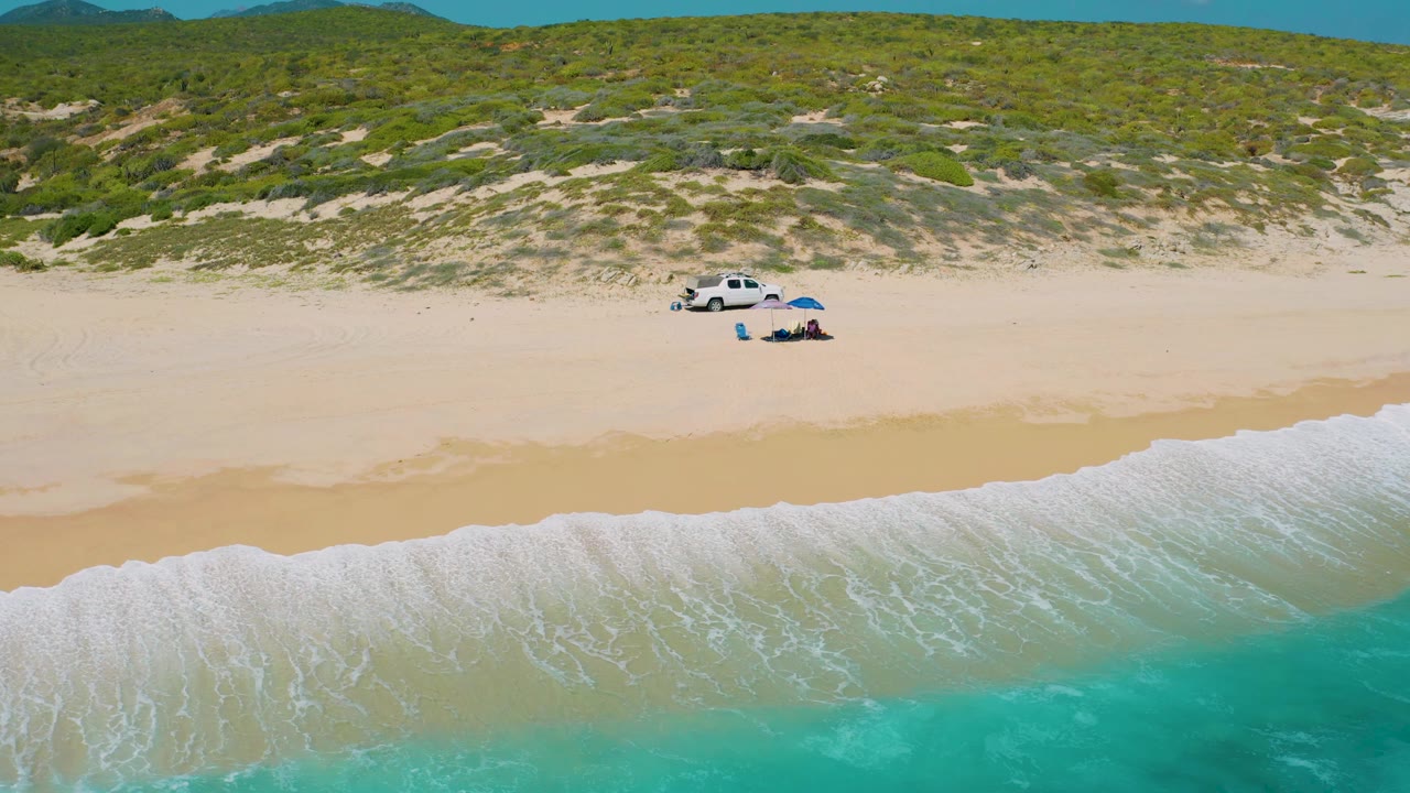 Drone frame over El Cardoncito beach with white surf entering from the lower right, a wide pale beach, and a white truck with a blue umbrella below the green dune line, with no visible people.