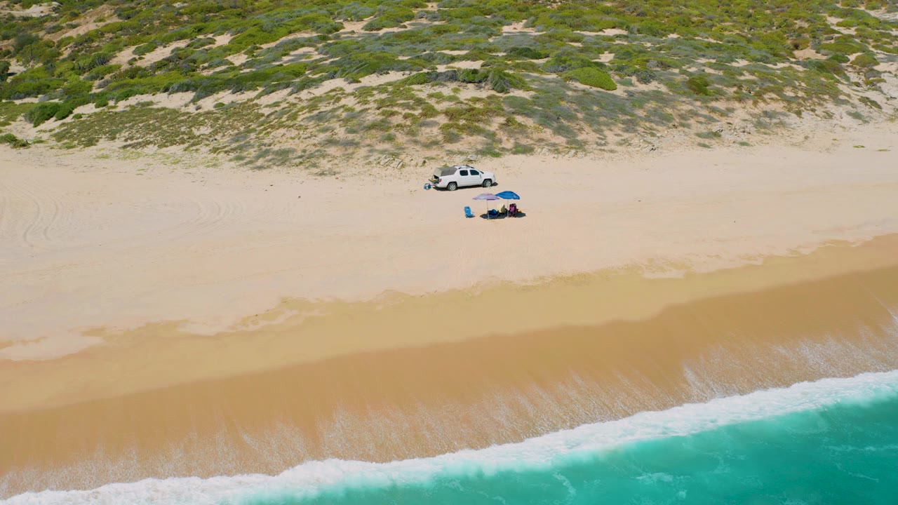 Opening aerial frame over El Cardoncito beach, Baja California Sur, with turquoise surf along the lower edge, pale sand in the center, low dunes above, a white vehicle, a blue umbrella, and no visible people.