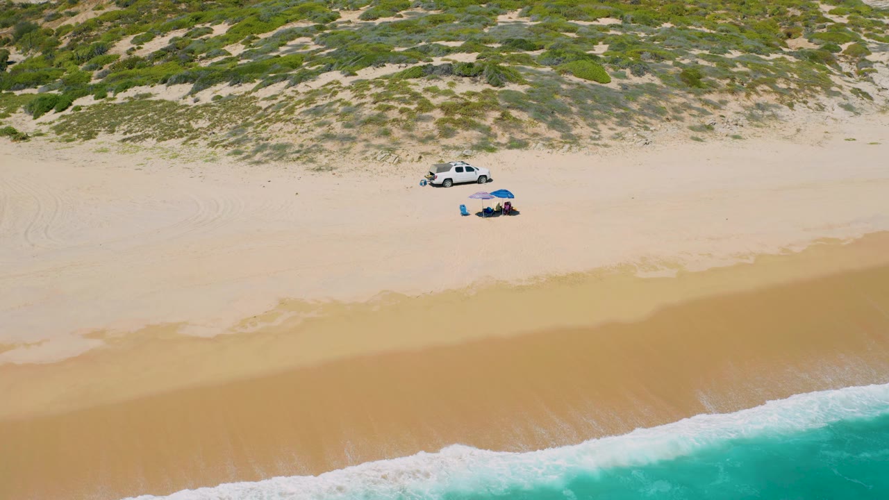 Aerial frame from El Cardoncito with the shoreline running diagonally across the lower portion, the parked white vehicle and blue umbrella near the dunes, and no visible people in the scene.
