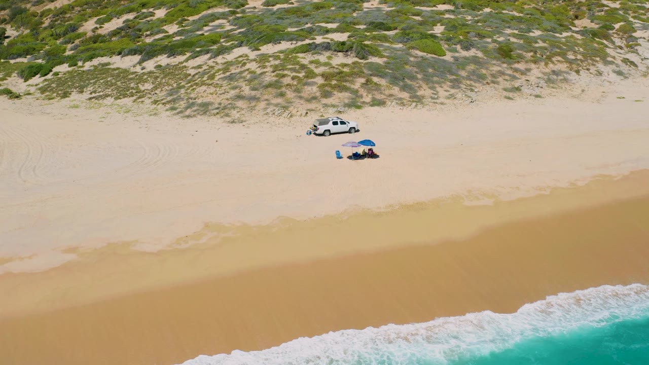Wide aerial beach frame with vehicle and umbrella above the surf line.