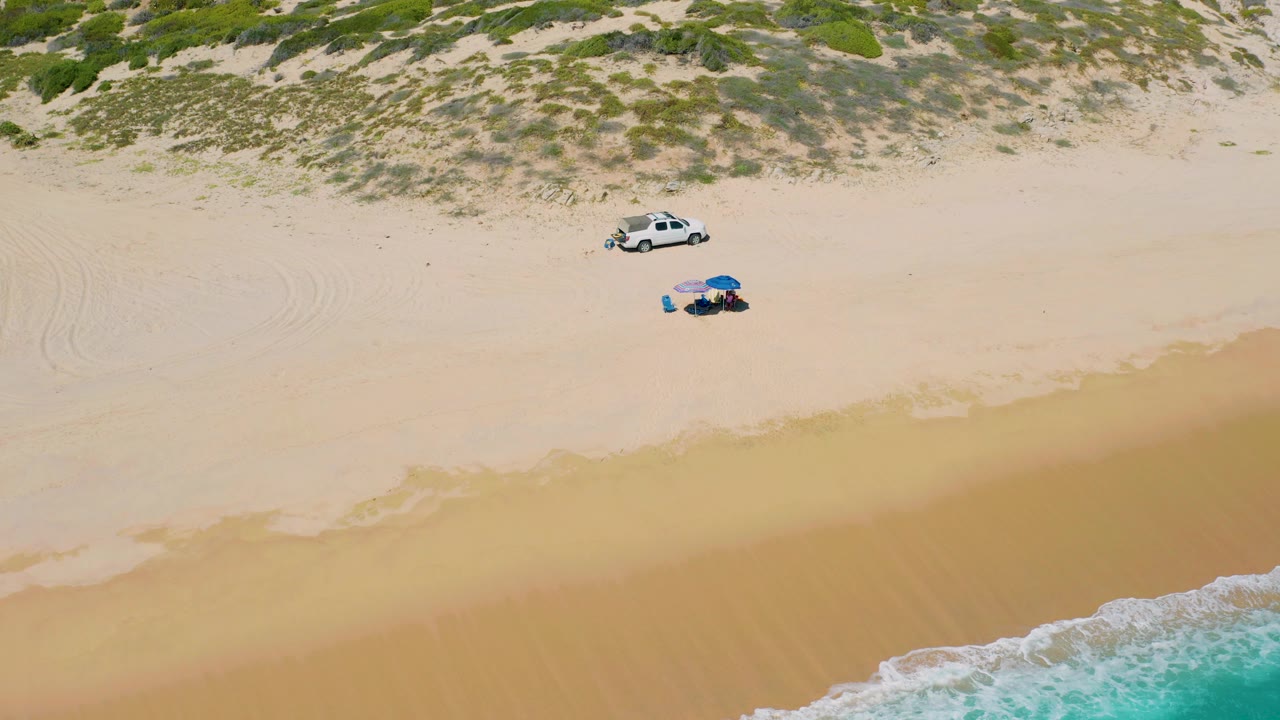 Overhead view in El Cardoncito, Baja California Sur, showing two people beneath a blue umbrella on pale sand, a white car parked near scrubby dunes, and surf at the lower edge.