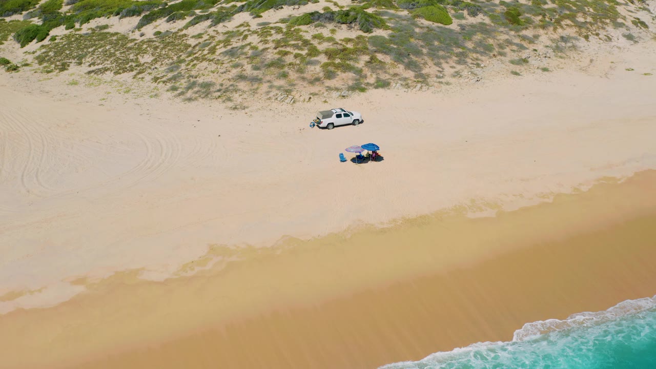 High overhead view in El Cardoncito showing the car slightly above the umbrella, two people on the sand, scrub vegetation along the upper frame, and turquoise water entering from the lower right.