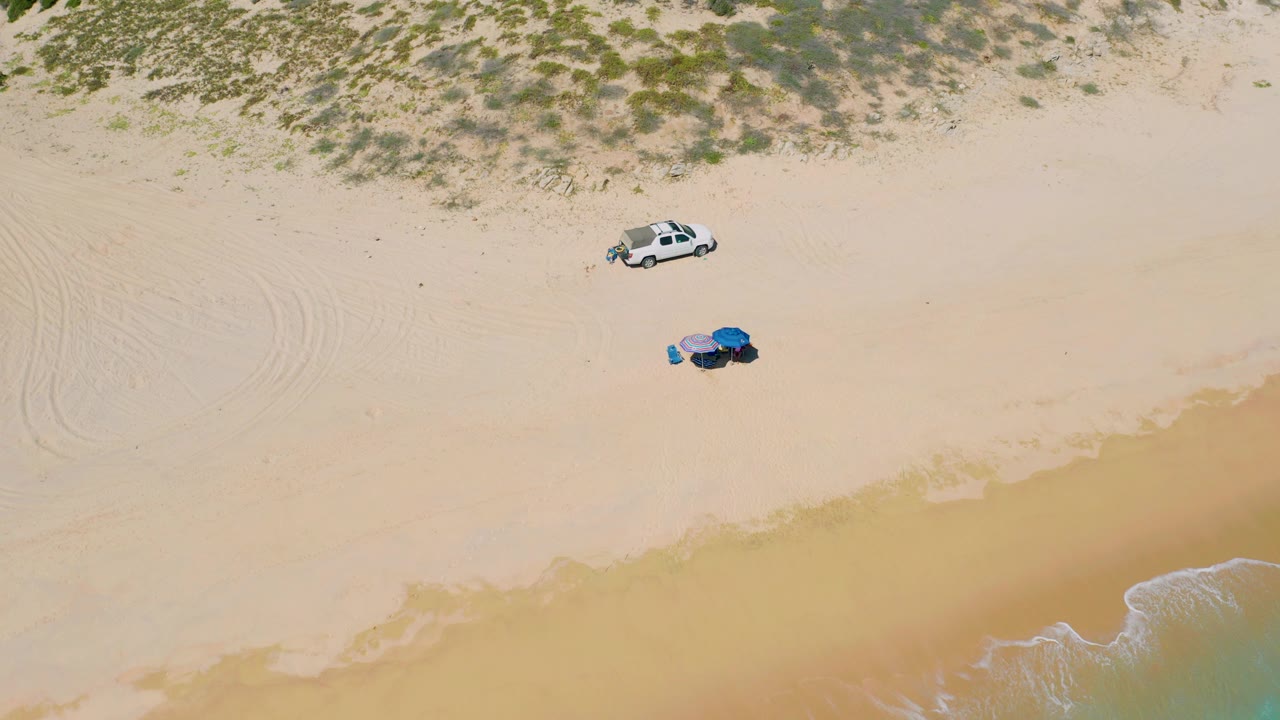Overhead frame from El Cardoncito, Baja California Sur showing a white vehicle near dune vegetation, blue and purple beach gear on pale sand, and a thin surf edge with no visible people.