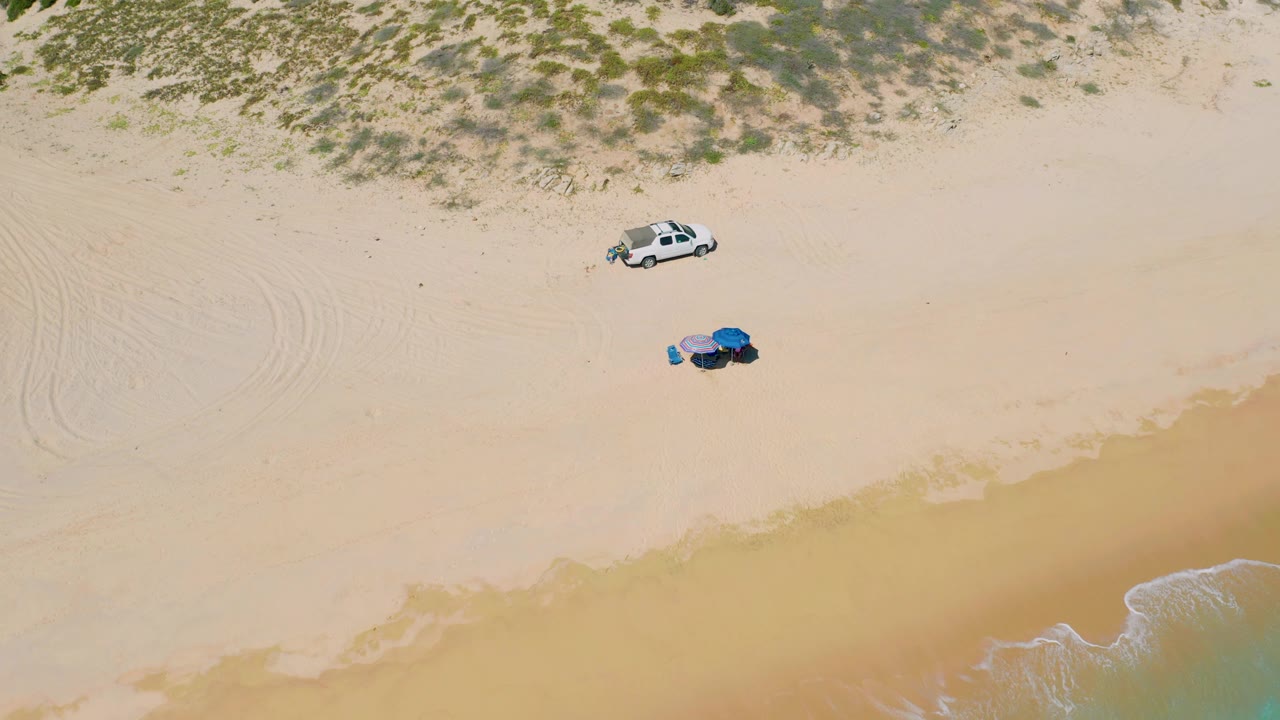 Aerial view over El Cardoncito beach with the parked white vehicle near the upper sand, clustered blue and purple beach gear below it, faint tire tracks, and no visible people.