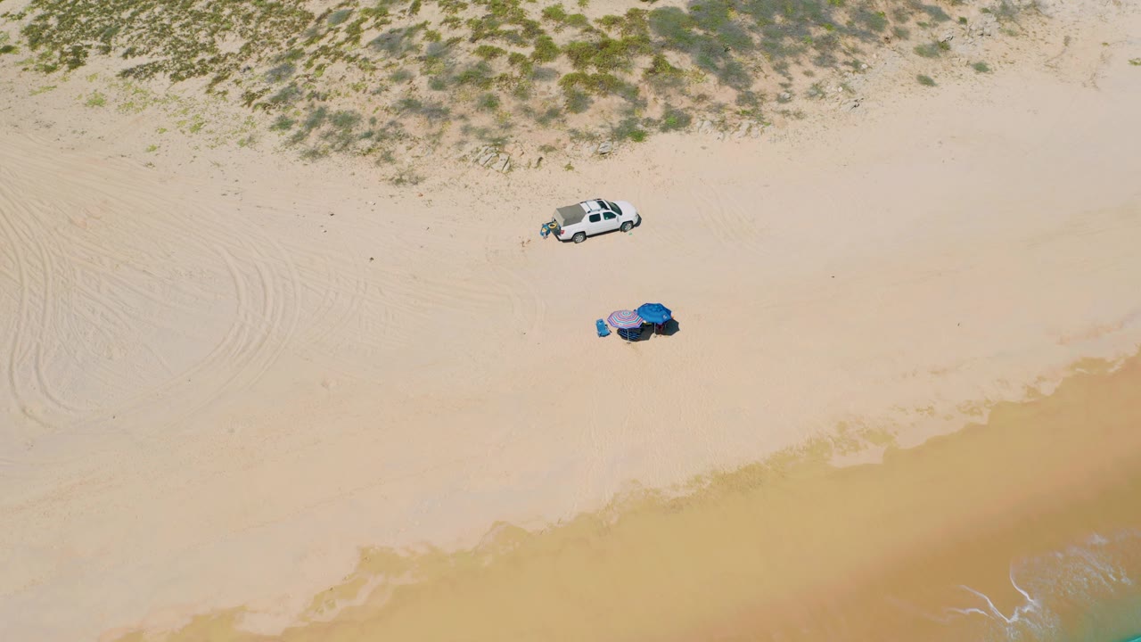 Top-down view at El Cardoncito with the white vehicle high in frame, two colored umbrella shapes on open sand, low dune scrub above, and a muted band of wave wash below with no visible people.