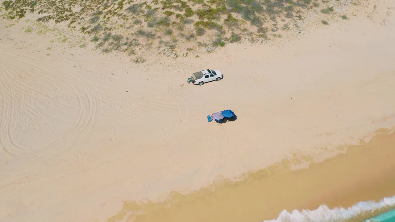 Overhead sand scene with tire tracks, parked vehicle, colored beach gear, and surf.