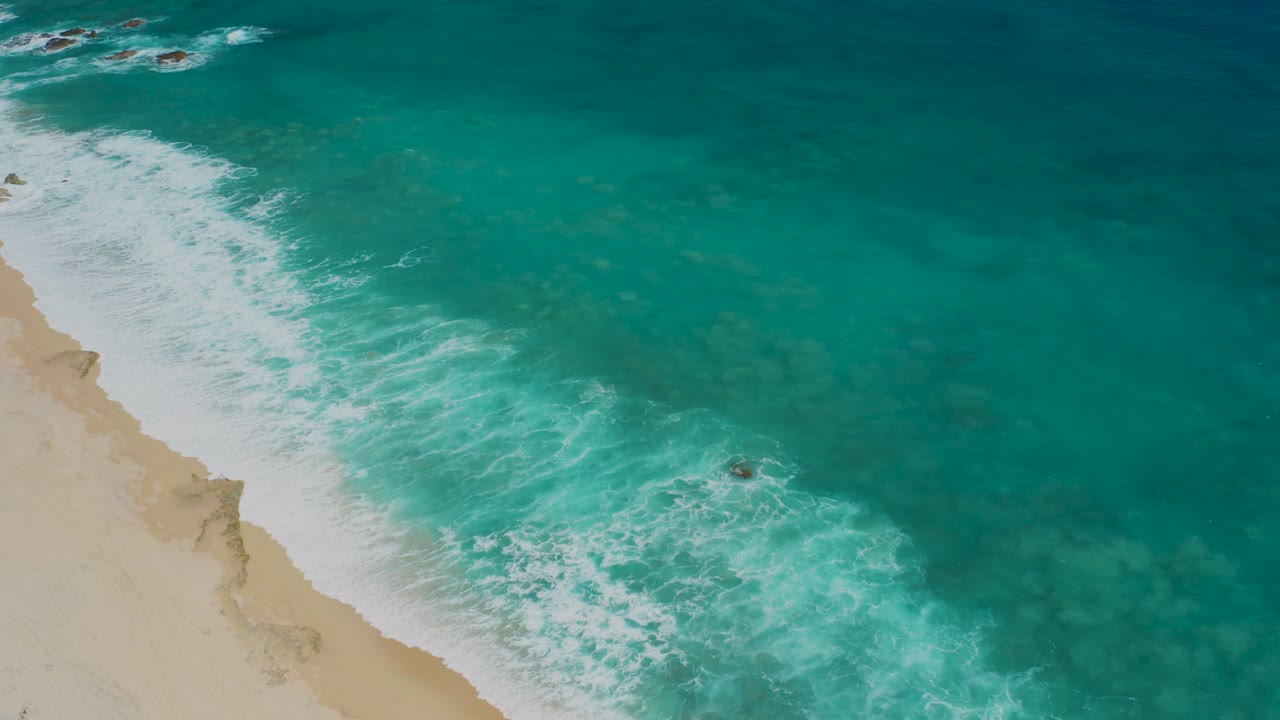 High aerial view at El Cardoncito showing pale sand on the left, turquoise water on the right, white foam along the shore, and a tiny person or dark figure near the surf.